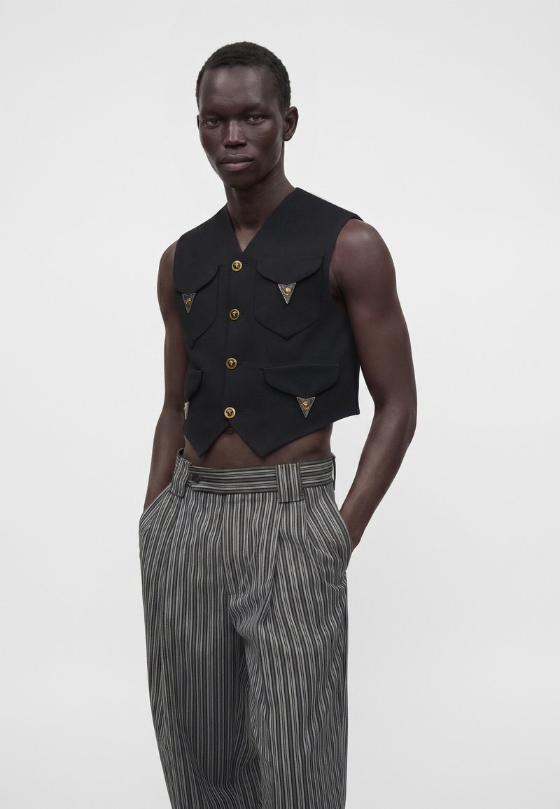 Homme mince à la peau foncée portant un gilet noir court sans manches avec des boutons dorés et un pantalon gris rayé taille haute, les mains dans les poches.