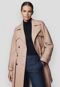 Beżowy wełniany płaszcz trench z podwójnym zapięciem na guziki, szerokimi klapami i paskiem w talii. Noszony na czarnym golfie i niebieskich spodniach.