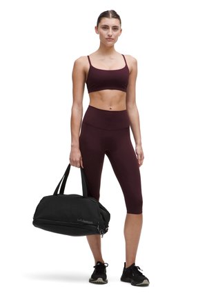 Sujetador deportivo burdeos y leggings de cintura alta hechos de tejido elástico, acompañados de un bolso de deporte negro con cremallera y logo.