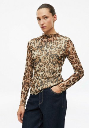 Woman wearing a long-sleeve leopard print top and dark blue jeans, standing with one hand in her pocket against a plain white background.