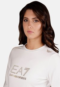 Weißes Langarmoberteil mit strukturiertem "EA7"-Logo und "EMPORIO ARMANI" in Gold, mit dekorativen Akzenten an der Schulter. Glatter Stoff.