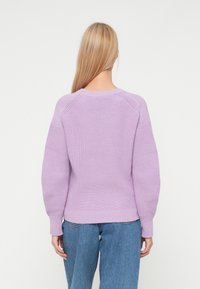 Mujer con cabello largo rubio, vistiendo un suéter de lana lavanda y jeans azules, de pie de espaldas contra un fondo blanco.