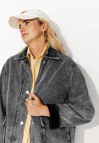 Femme aux cheveux clairs portant une casquette blanche, des boucles d'oreilles en or, une chemise à rayures jaunes et une veste en denim noire, regardant légèrement vers la gauche.