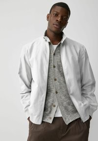 Chaqueta gris claro con cremallera y cuello alto, usada sobre un suéter de punto gris y una camisa blanca; presenta un corte relajado y bolsillos laterales.