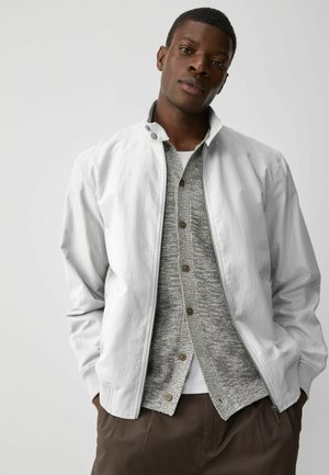 Chaqueta gris claro con cremallera y cuello alto, usada sobre un suéter de punto gris y una camisa blanca; presenta un corte relajado y bolsillos laterales.