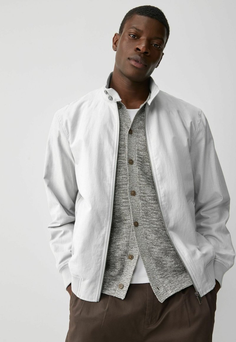 Chaqueta gris claro con cremallera y cuello alto, usada sobre un suéter de punto gris y una camisa blanca; presenta un corte relajado y bolsillos laterales.