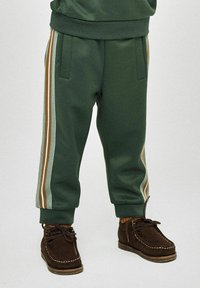 Pantalons de survêtement verts avec des jambes fuselées, présentant des bandes contrastantes marron et bleu clair sur les côtés ; accompagnés de chaussures en daim marron.