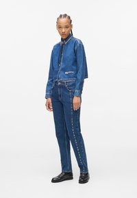 Denim jack en broek in donkerblauw. De jack heeft studs op de schouders en een cropped ontwerp. De broek heeft studs aan de zijkanten. Het model draagt zwarte schoenen.