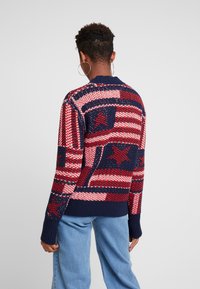 Persoon met kort, krullend haar draagt grote oorbellen en een gebreide trui in rood, wit en blauw met sterrenpatronen, gecombineerd met lichtblauwe spijkerbroek.