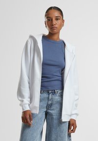 Felpa bianca con zip e texture morbida, con un cappuccio ampio e tasche frontali. Indossata sopra un top a coste blu e jeans in denim chiaro.