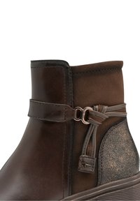 Bottine marron avec textures lisses et en daim, bride décorative avec anneau en métal et pompons en cuir noués, et panneau de talon pailleté.