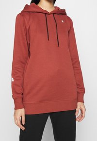 Röd huvtröja med svarta snoddar, ribbade muddar och nederkant. Har en liten logotyp på bröstet och vit text på ärmen.