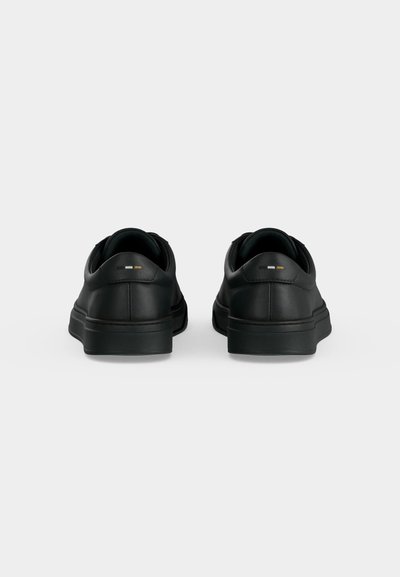 Paire de baskets noires basses vues de dos, sur un fond gris clair uni.
