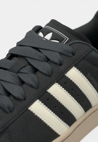 Černé tenisky se šedými tkaničkami, semišovým svrškem, třemi krémovými pruhy a gumovou podešví. Na jazyku je nášivka s logem.