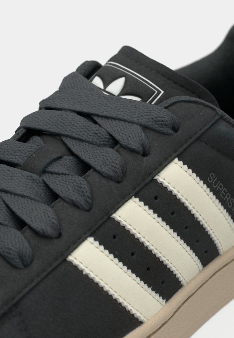 Svarta sneakers med gråa snören, ovandel i mocka, tre krämfärgade ränder och en gummisula. Har en logotypmärke på tungan.