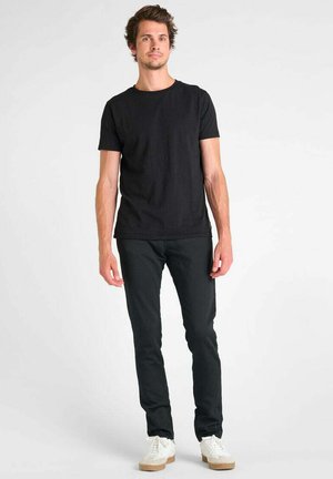 Man staat tegen een witte achtergrond, draagt een zwart T-shirt, zwarte broek en witte sneakers, kijkt met een neutrale uitdrukking vooruit.