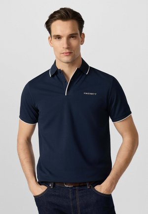 Jonge man die een marineblauw Hackett poloshirt met witte bies draagt, staat met zijn handen in zijn zakken tegen een effen grijze achtergrond.