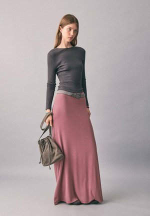 Femme jeune portant un haut gris ajusté à manches longues et une jupe maxi rose taille haute, tenant un sac à main en suede gris à ses côtés.