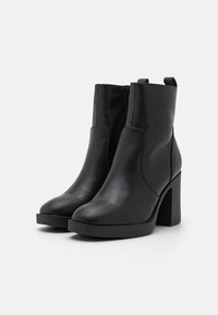 Bottes noires à talons pour cheville en cuir synthétique lisse. Dotées d'une fermeture éclair latérale et d'un talon rectangulaire épais. Design minimaliste.