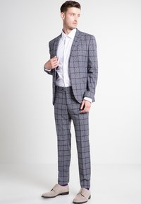 Grijze geruite pak met donkerrode en marineblauwe accenten, gecombineerd met een wit shirt en lichtgrijze schoenen. Taillering met zichtbare texturen.