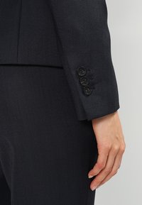 Marinblå kavaj i strukturerad tyg med skräddarsydd passform, skarpa slag och fyra svarta knappar på ärmen för säker stängning.