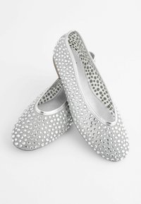 Ballerines argentées ornées de strass ronds uniformément espacés, une chaussure posée à l’intérieur de l’autre sur un fond blanc.