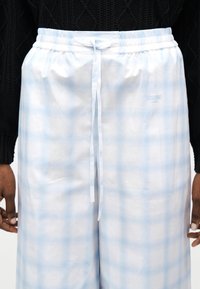 Ανδρικό ανοιχτό μπλε και λευκό καρό παντελόνι με κορδόνι και λογότυπο Moschino Jeans στο δεξί μηρό, συνδυασμένο με μαύρο πλεκτό πουλόβερ.