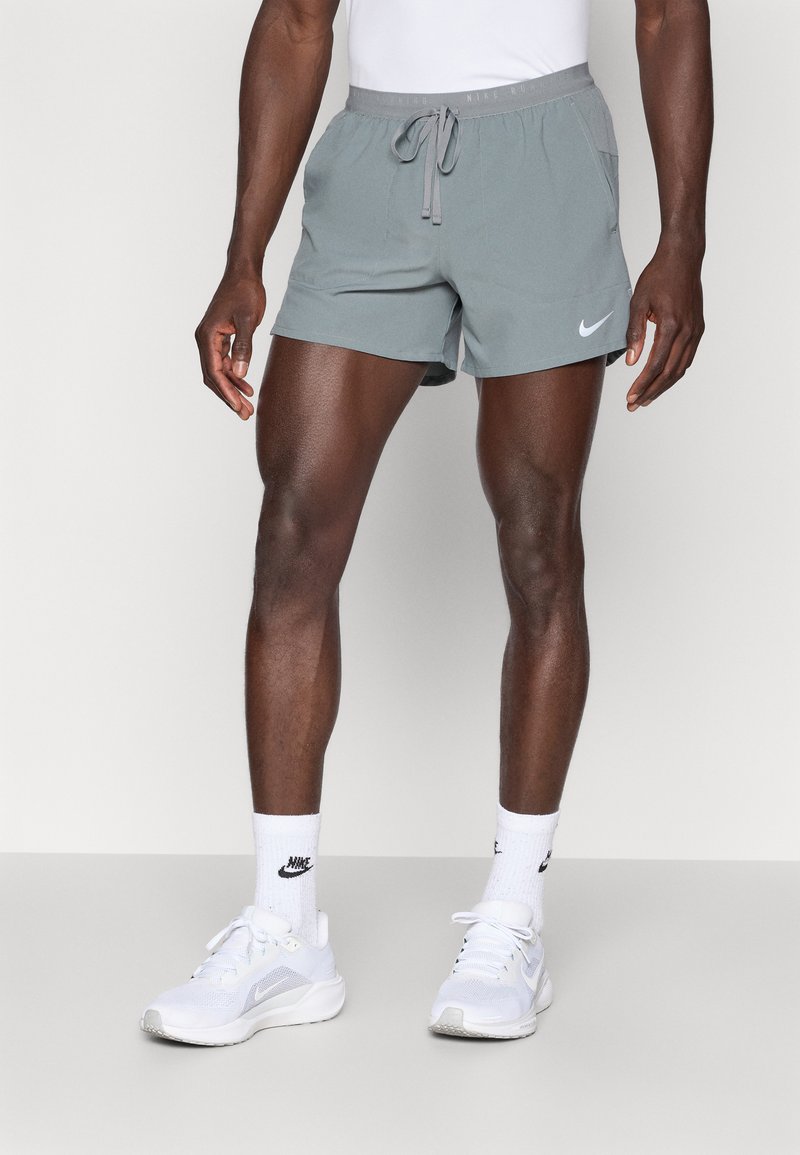 Αθλητικό σορτς σε ανοιχτό γκρι χρώμα με ελαστική μέση και κορδόνι, πλαϊνές τσέπες και λευκό λογότυπο Nike. Φοριέται με λευκά αθλητικά παπούτσια και κάλτσες με σχέδια στο μήκος του αστραγάλου.