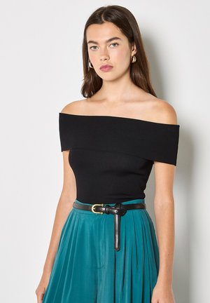 Jeune femme aux longs cheveux bruns portant un haut noir décolleté, une jupe plissée bleu sarcelle, une ceinture noire et des boucles d'oreilles créoles dorées sur fond blanc.