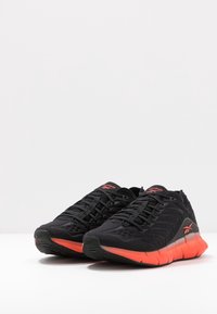 Chaussures de sport noires avec une tige texturée, dotées d'une semelle et d'accents orange, d'un bout rond et d'un design classique à lacets.