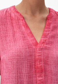 Blouse rose, texturée, avec un design en V et un motif froissé subtil. Manches courtes et une fermeture à bouton simple à l'avant.