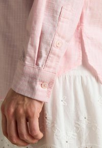 Svetloružová košeľa na gombíky s károvaným vzorom, s vyšitým logom na manžete a gombíkmi zodpovedajúcimi farbe látky.