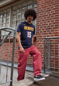 Jūras spēku t-krekls ar uzrakstu "DENVER" un numuru "15" dzeltenā un baltā krāsā, saskaņots ar brīviem sarkaniem biksēm un melniem sporta apaviem, stāv uz kāpnēm.