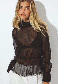 Sort, gennemsigtig bluse med plisseret tekstur, høj hals og flæse forneden. Designet inkluderer en tætsiddende talje og lange, svajende ærmer.