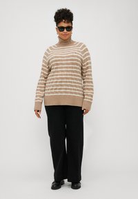 Pulover bej în dungi cu guler înalt, având un tricot texturat și manșete cu nervuri, purtat împreună cu pantaloni negri largi și pantofi negri.