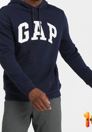Person iført en marineblå GAP-hoodie og grå bukser, stående med armene let udstrakte mod en ensartet hvid baggrund.