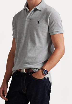 Man draagt een lichtgrijs poloshirt met korte mouwen en een klein paardenlogo, donkere jeans, een bruine riem en een polshorloge met een blauwe wijzerplaat en een leren band.