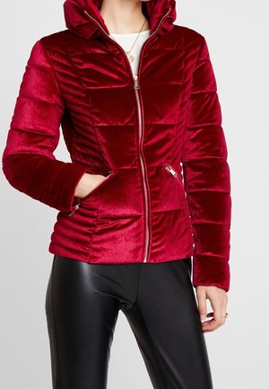 Femme portant une veste matelassée ajustée en velours rouge avec une fermeture éclair et des poches, associée à un pantalon en cuir noir et un haut blanc.