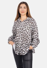 Leopardmuster-Hemd mit Knopfverschluss aus leichtem Stoff, mit Kragen, langen Ärmeln und Fronttaschen, kombiniert mit schwarzen, enganliegenden Hosen.