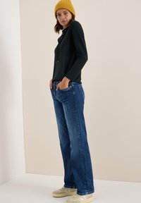 Dunkles Langarmoberteil mit Kordeln, kombiniert mit locker sitzenden blauen Jeans und beigen Sneakers, vor einem neutralen Hintergrund.