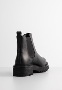 Bottes Chelsea en cuir noir avec panneaux latéraux élastiques, talon surélevé et semelle extérieure en caoutchouc robuste. Texture lisse et détails de couture minimaux.