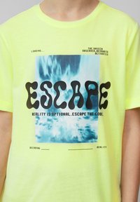 Neon gele T-shirt met een blauw en witte abstracte graphic en de tekst "ESCAPE" en aanvullende zinnen in het zwart. Katoenen materiaal.