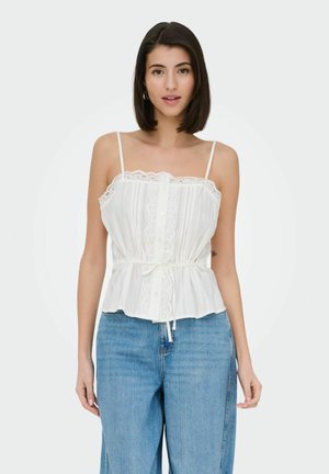 Mladá žena oblečená v biele tielko bez rukávov s čipkou a modré rifle s vysokým pásom, stojaca vzpriamene pred jednofarebným pozadím.