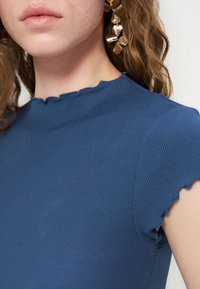 Gros plan d’une femme portant un haut bleu côtelé avec un col et des manches à bord ondulé, montrant des boucles d’oreilles florales dorées et les cheveux détachés.