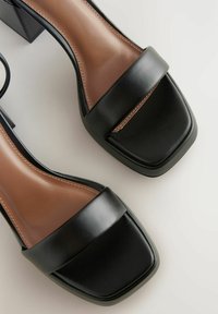 Černé kožené sandály na podpatku s širokým páskem a čtveratou špičkou. Béžové vnitřní podšívání s kontrastním prošíváním. Design s blokovým podpatkem.