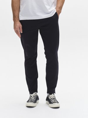 Svarta avsmalnande byxor med slät textur, två sidofickor och ribbade muddar, kombinerade med svartvita sneakers.