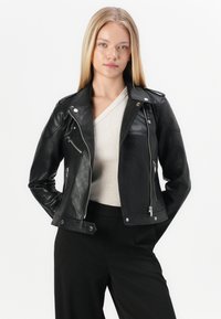 Svart skinnmotorjakke med sølvfarget hardware, med glidelåslommer og en tettsittende design. Båret over en lys beige topp.
