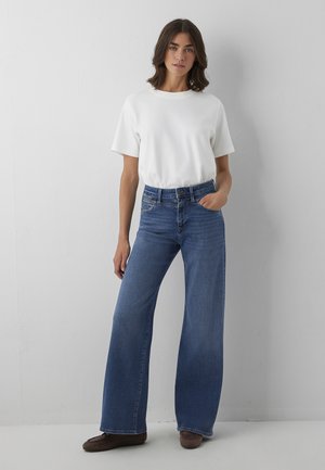 Frau steht an einer weißen Wand, trägt ein einfaches weißes T-Shirt, hoch taillierte, weite blaue Jeans und braune Schuhe, mit einer Hand in der Tasche.