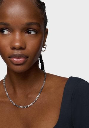 Giovane donna con capelli intrecciati che indossa orecchini a cerchio d'argento, una collana floreale d'argento e una maglietta scura con scollo a barca, che guarda di lato.