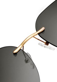 Černé sluneční brýle s přehnanými čočkami, zlatý kovový most a průhledné nosní podložky. Mají elegantní, zakřivený design a minimalistické akcenty.